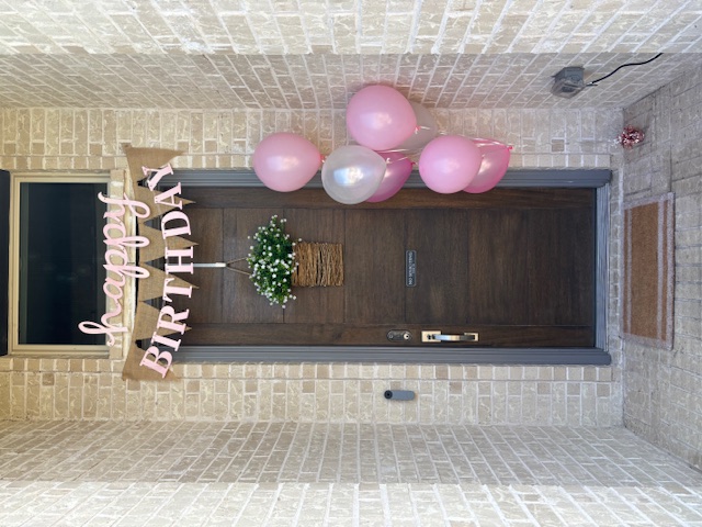 Festive birthday door decoration with happy birthday banner, wicker basket of white flowers, and pink and silver balloons on brick entrance