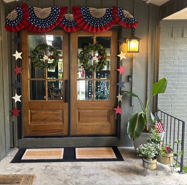 Elegant wooden double doors with patriotic decorations featuring red, white, and blue paper fan bunting, green wreaths, star garlands, and lantern lighting